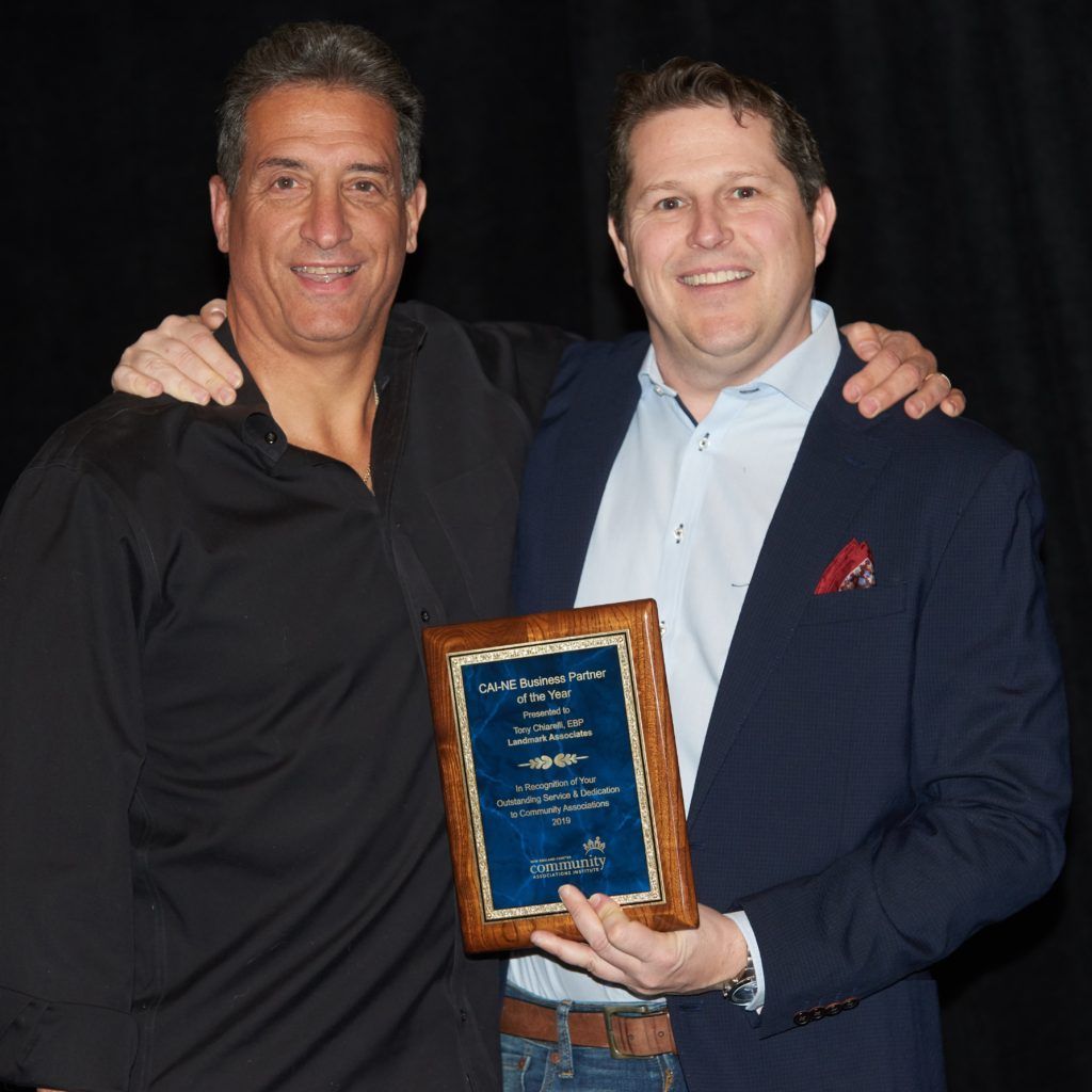 Two men with plaque, one in suit, other in black shirt, both smiling, indoor setting.