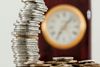 Stack of coins in front of a blurred clock. Focus is on the silver coins, hinting at wealth and time.