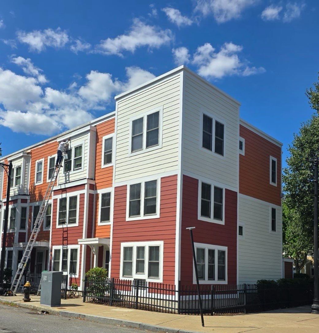 Three-story building with red, orange, and white siding. Two people on a ladder, sunny day, blue sky.