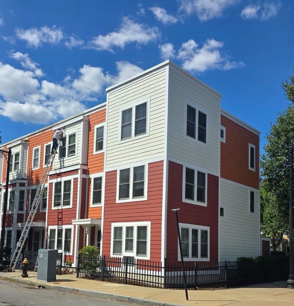 Three-story building with red, orange, and white siding. Two people on a ladder, sunny day, blue sky.