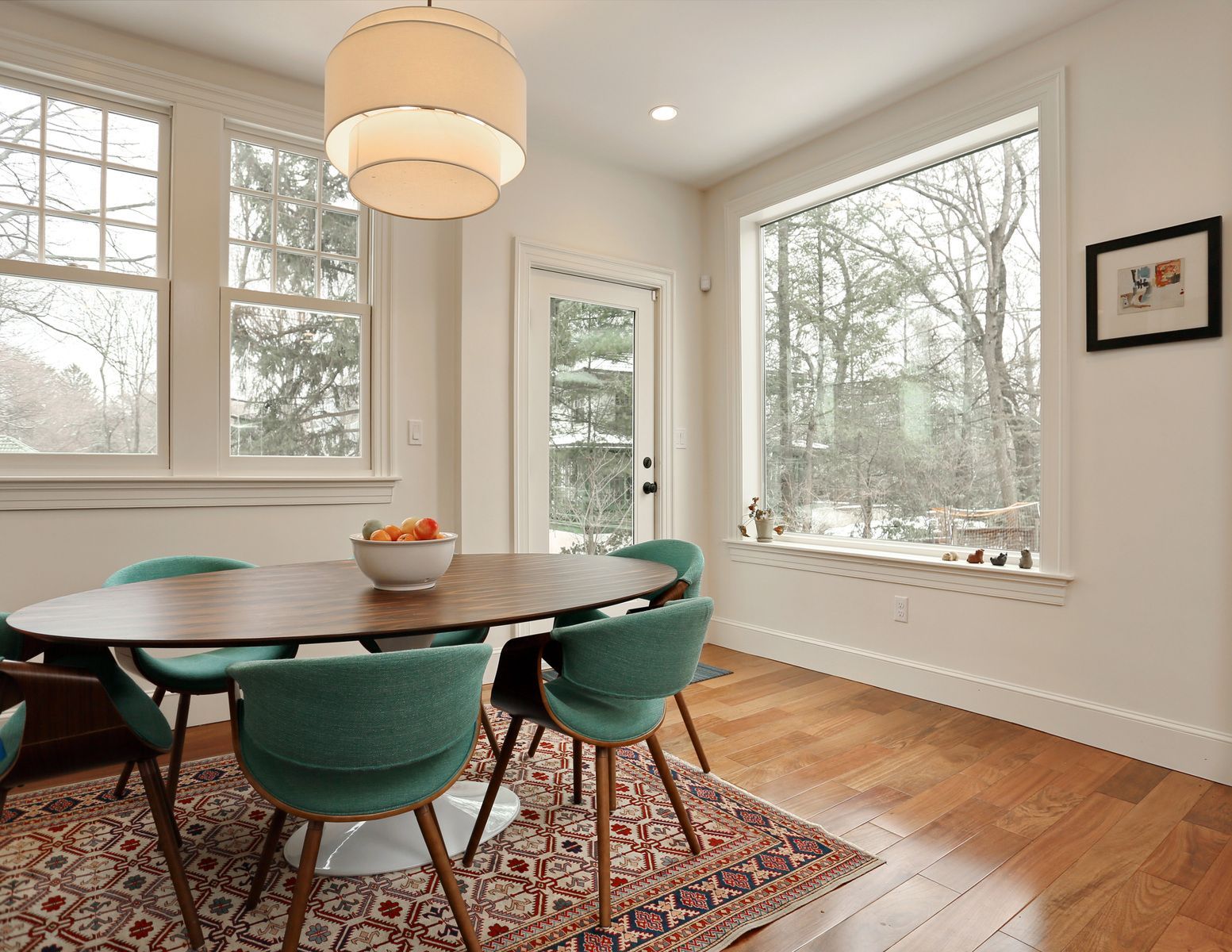 Dining room with a wooden table, teal chairs, and large windows overlooking trees.