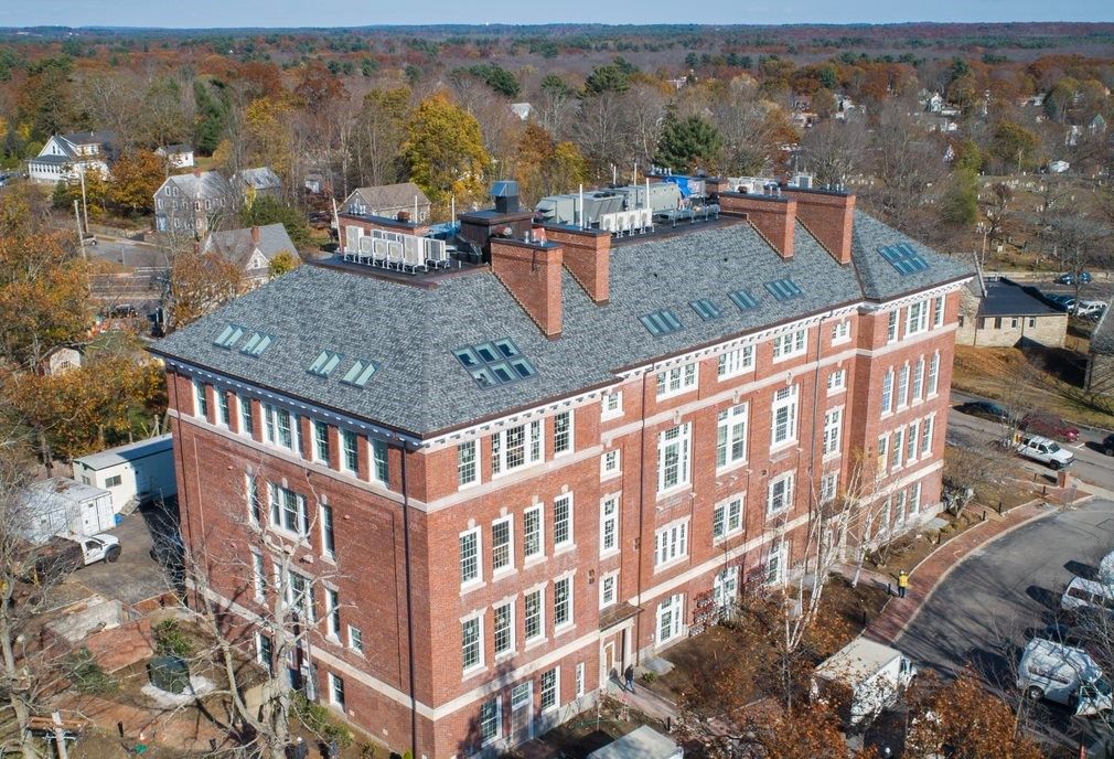 Aerial view of a four-story brick building with a dark gray roof and several windows, surrounded by trees.