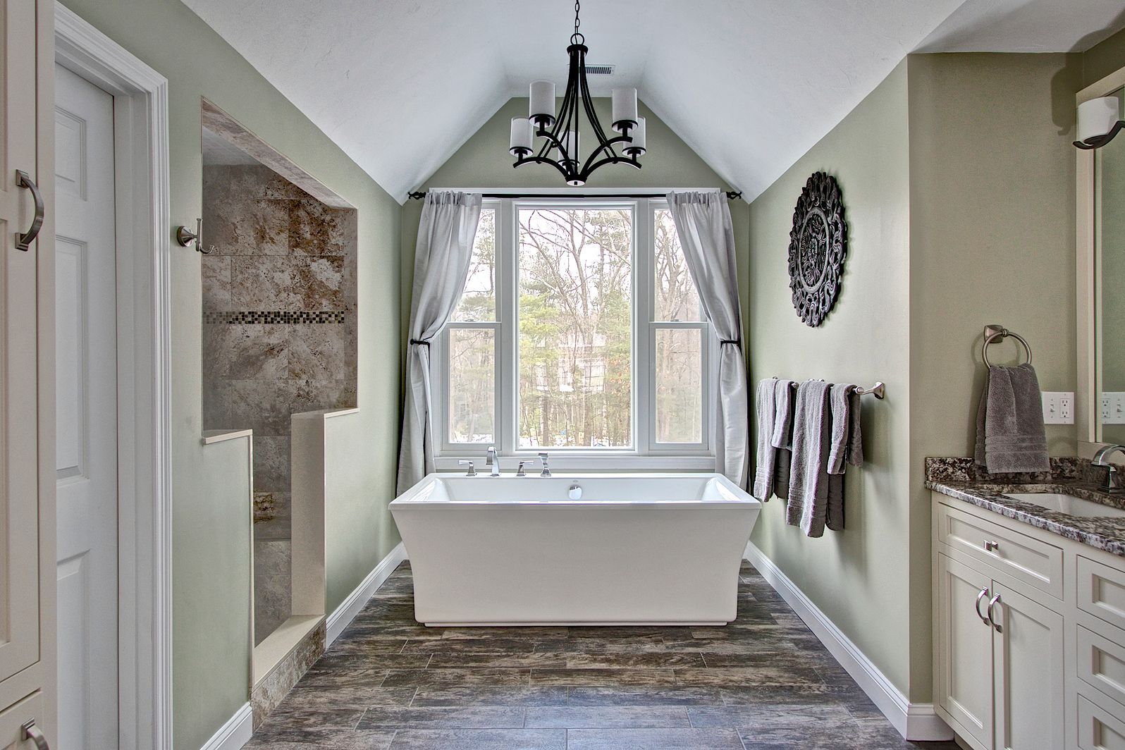 Bathroom with a white freestanding tub under a window, gray walls, and a hanging chandelier.
