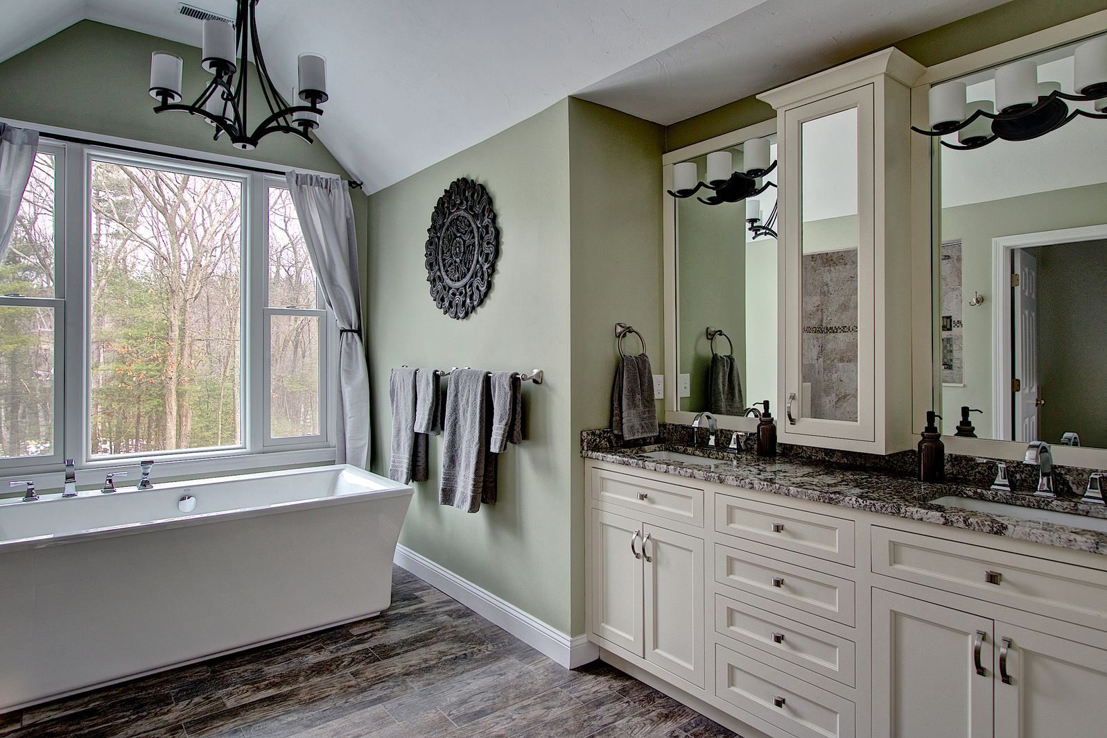 Spacious bathroom with soaking tub by a window, double vanity, green walls, and ornate black chandelier.