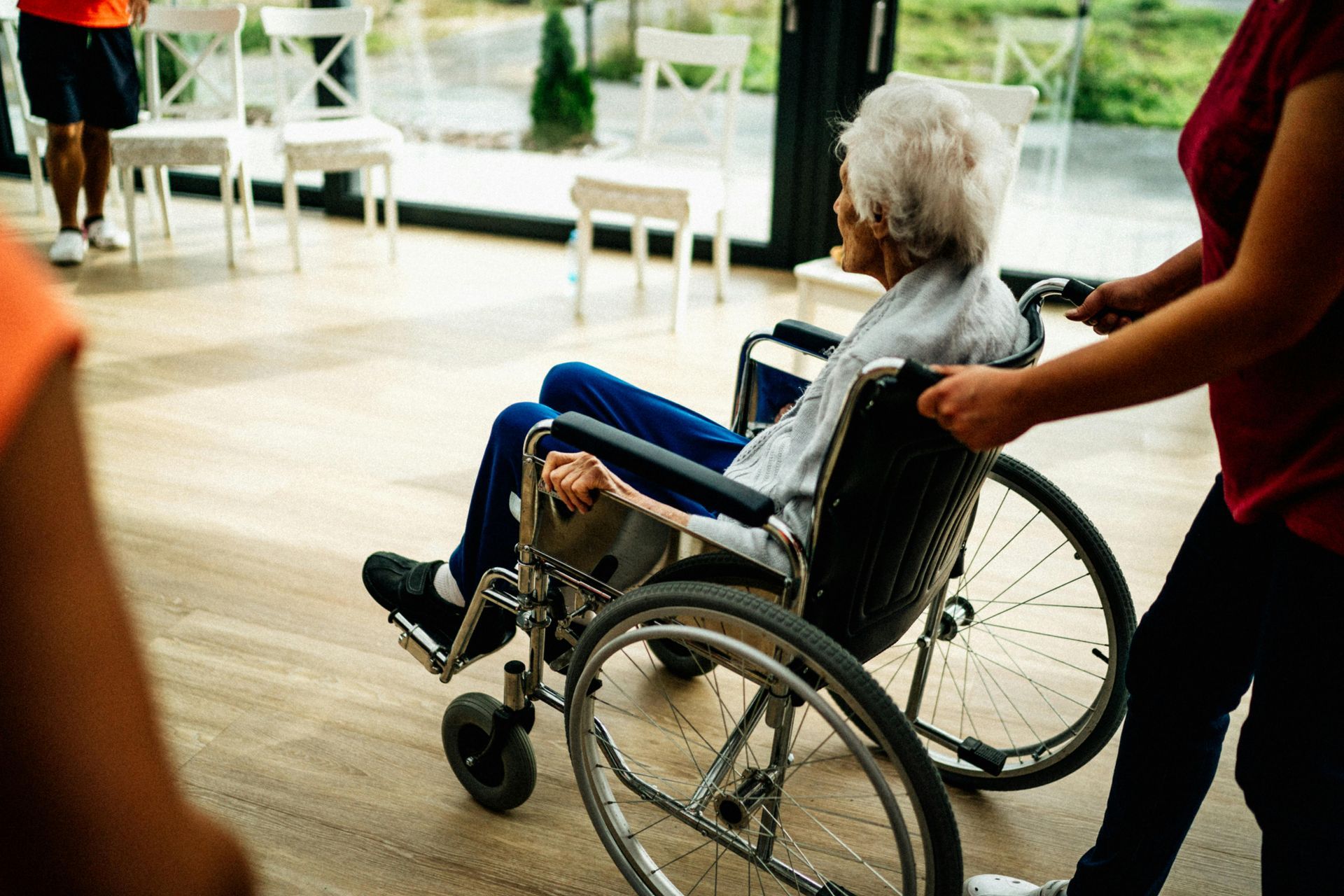 Person in wheelchair being pushed by another person indoors, near chairs and windows.