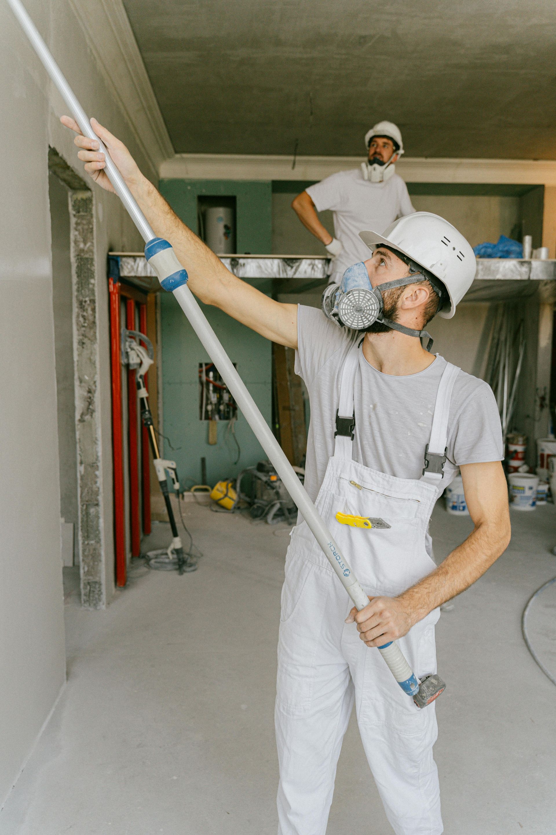 Two construction workers plastering a wall in a room. One uses a long-handled tool; the other observes.
