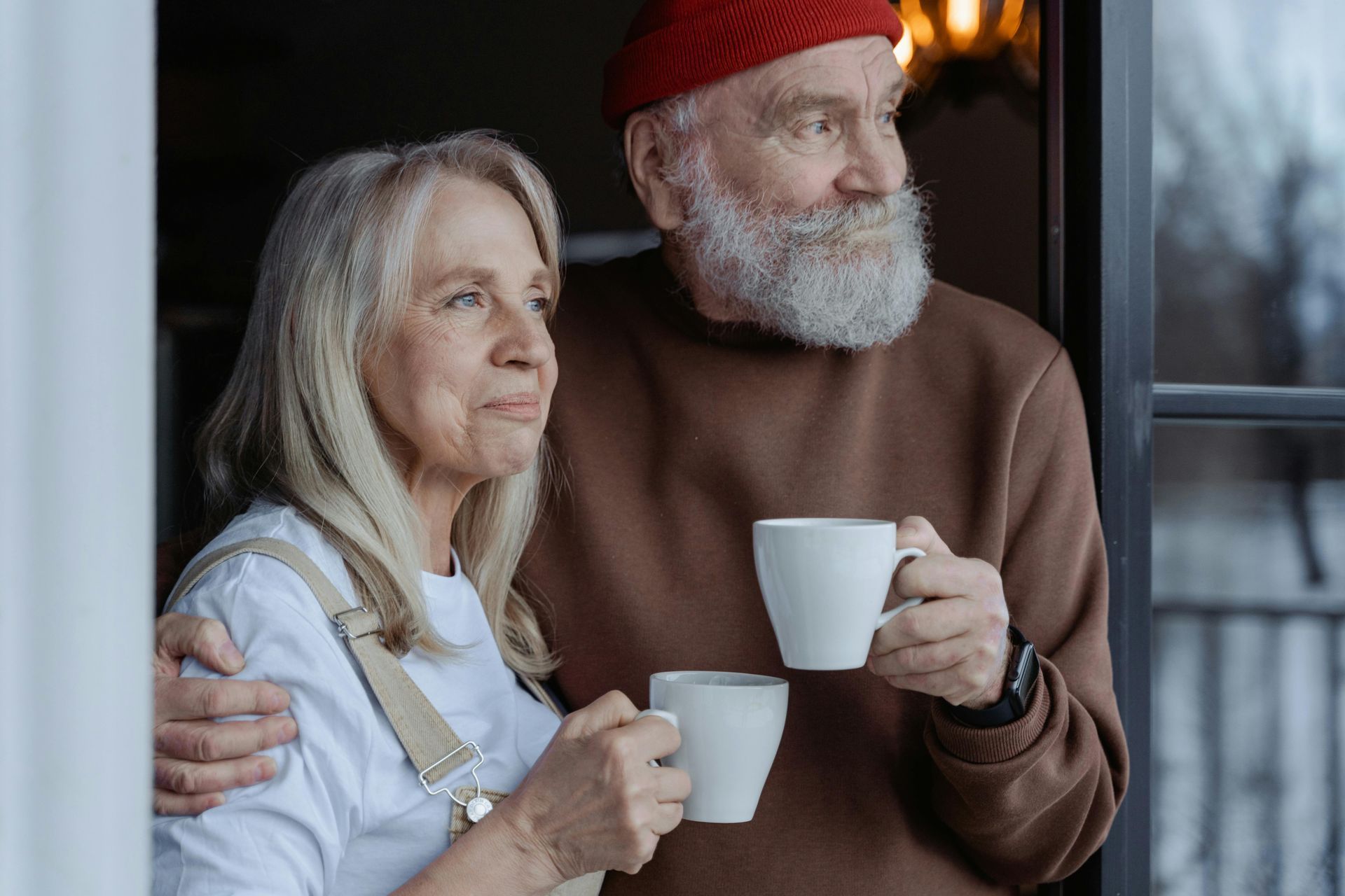Older couple holding mugs, looking out window. Man in red hat, brown sweater; woman in white shirt, overalls.