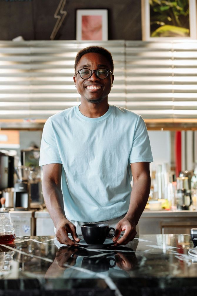 Man holding a black coffee cup, smiling at the camera, inside a cafe.