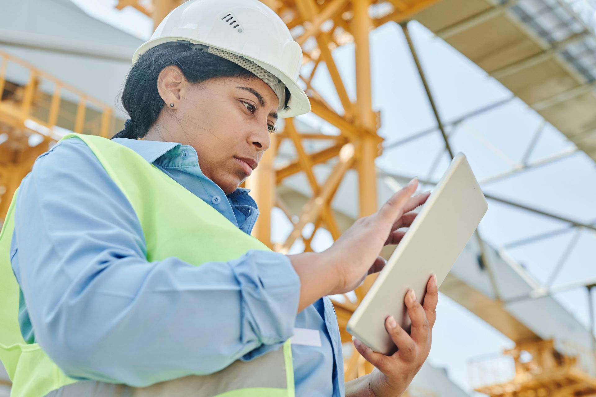 Woman in hard hat and safety vest, looking at tablet, construction site background.
