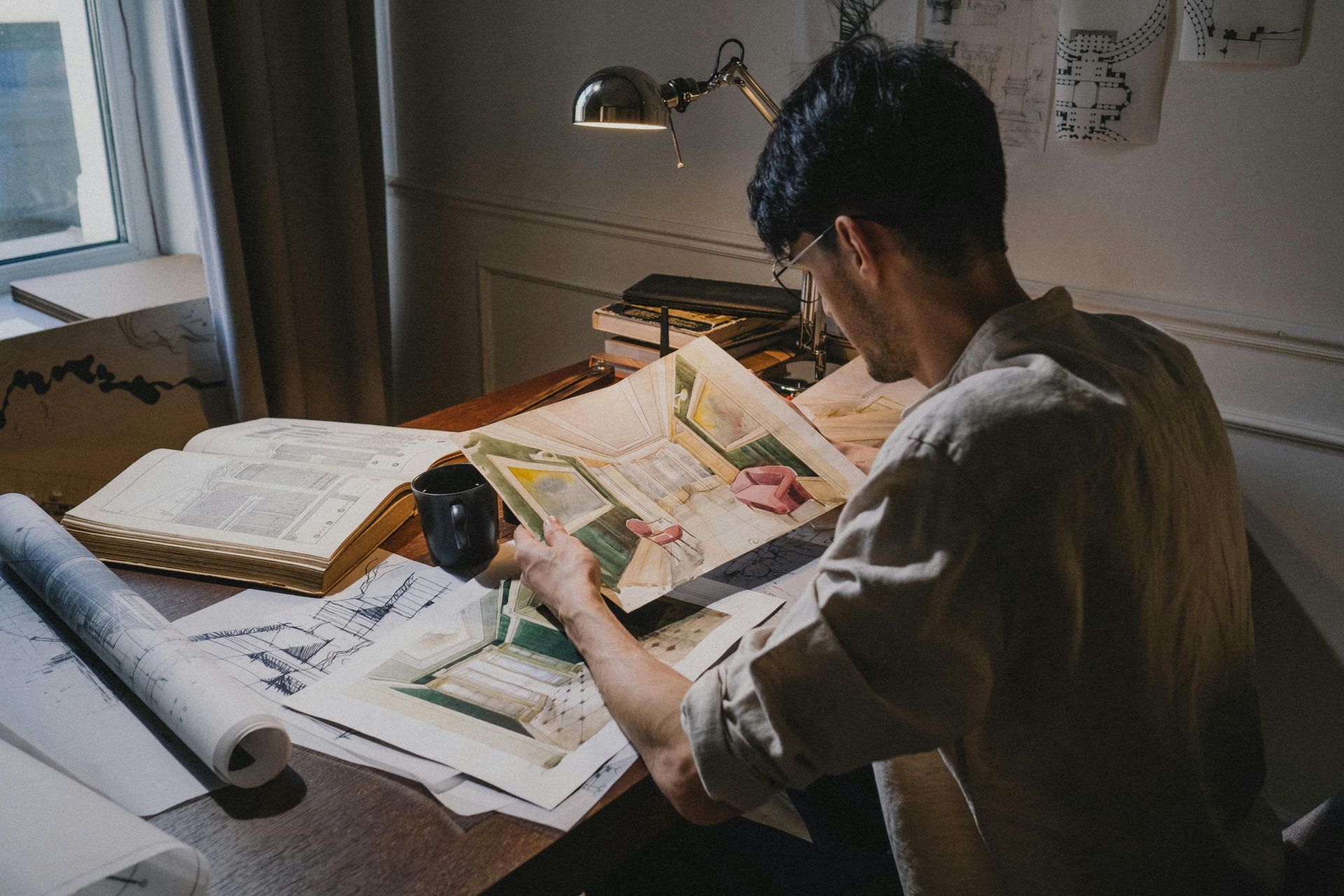 Person reading large document at desk with lamp, books, and blueprints.