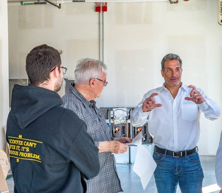 Four men in a light-filled room, one gesturing with hands, discussing papers near metal equipment.