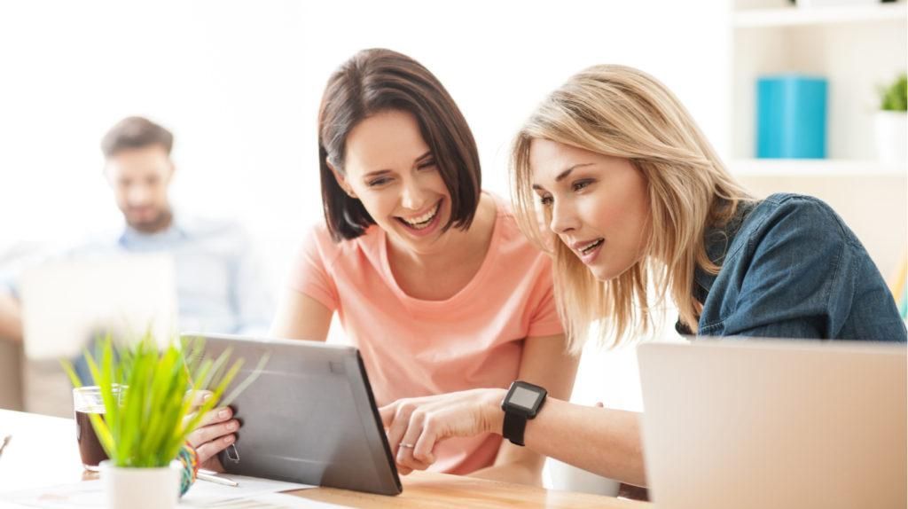 Two women are sitting at a table looking at a tablet computer.