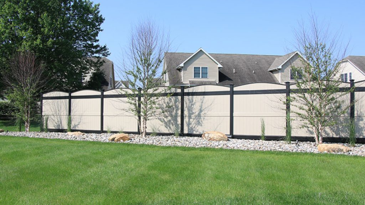 Beige fence with black frame in front of a house, trees, and green grass on a sunny day.