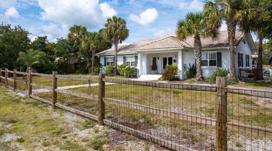 A white, single-story house with a tiled roof behind a wooden wire fence, surrounded by palm trees and green bushes.