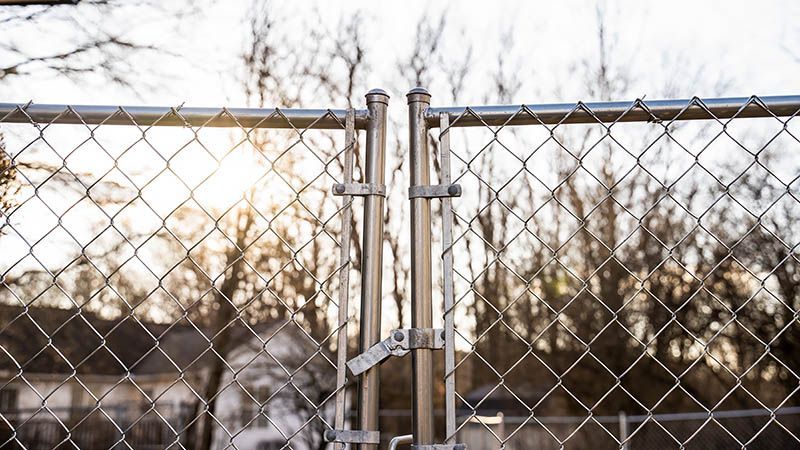 A chain link fence with a gate open and trees in the background.