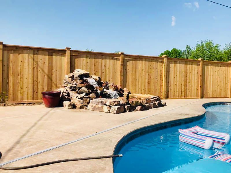 A wooden fence surrounds a swimming pool with a waterfall in the background.