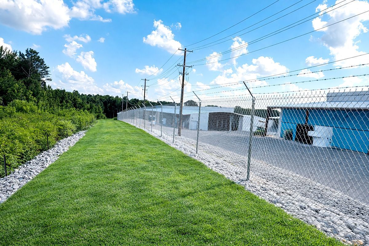A chain-link fence topped with barbed wire runs alongside a green lawn, gravel, and a blue industrial building under clouds.