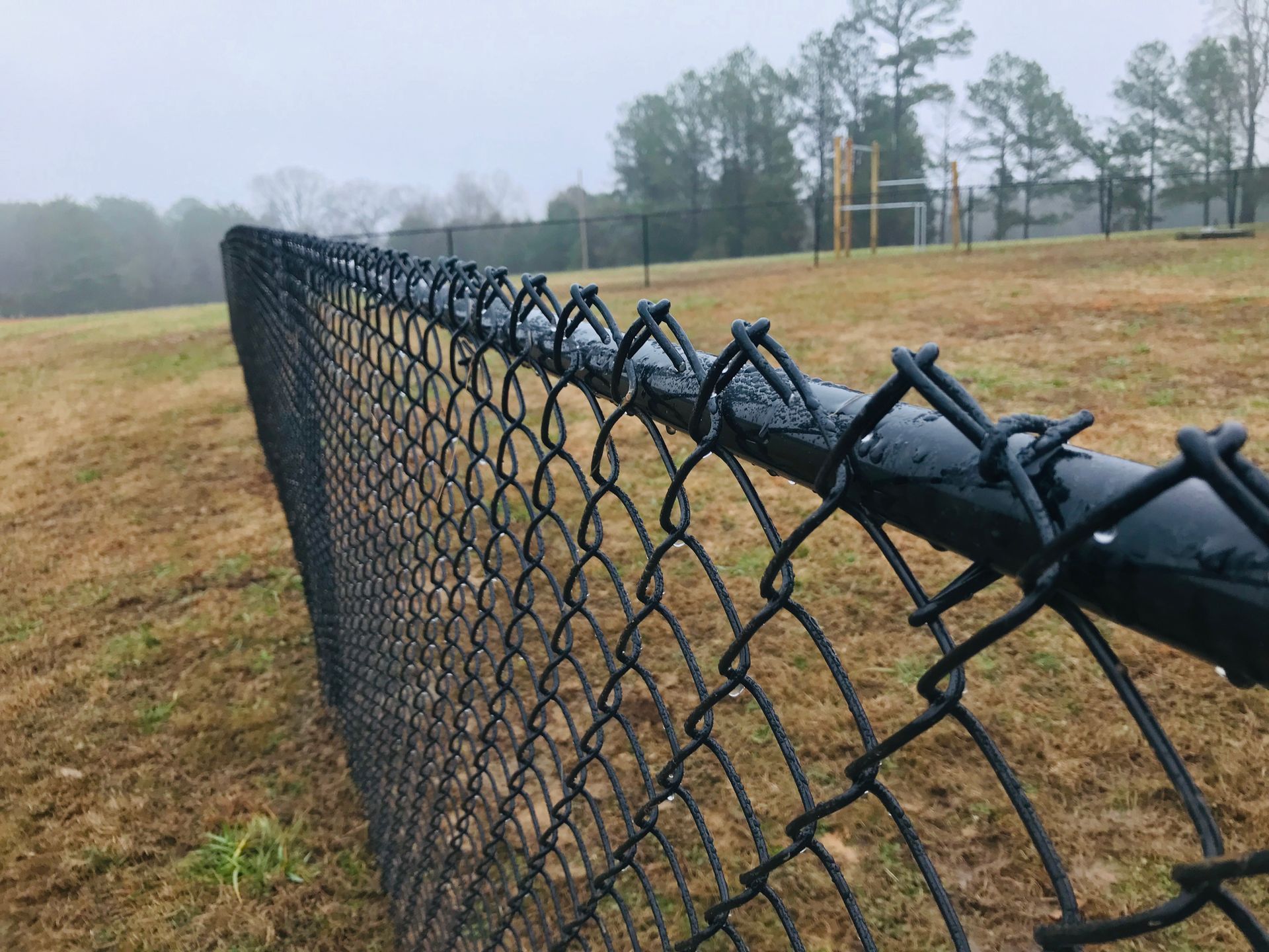A close-up, angled view of a black chain-link fence on a grassy field under a cloudy, overcast sky.