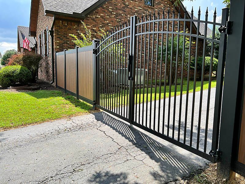 A black metal gate is open to a driveway in front of a brick house.