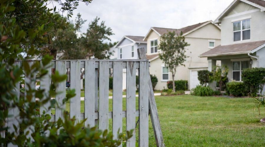 A suburban street with light-colored houses, green lawns, and a wooden picket fence in the foreground.