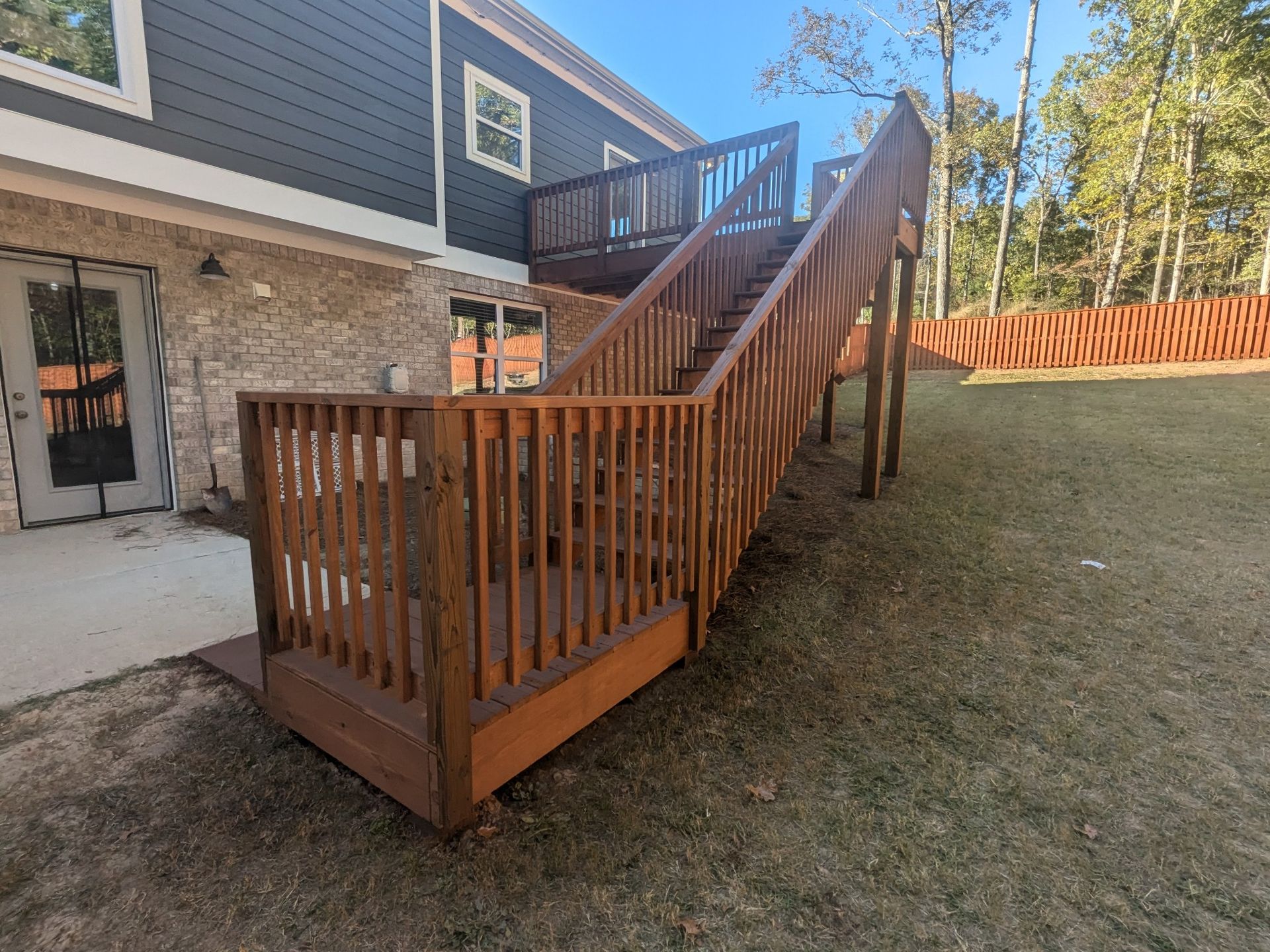 A wooden deck with a staircase leading from a second-story balcony down to a paved patio area in a backyard.