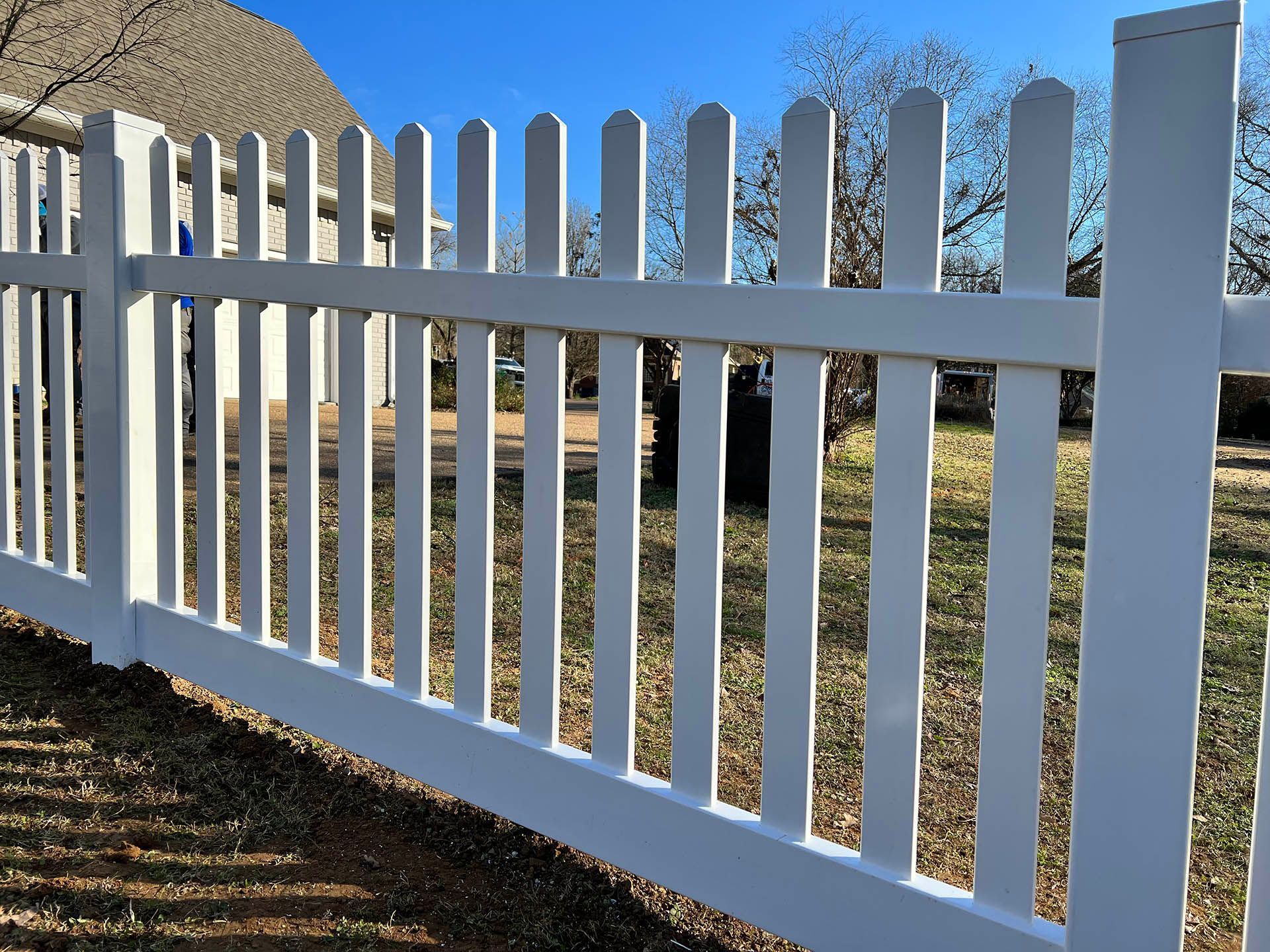 White picket fence in a yard with a house visible in the background on a sunny day.