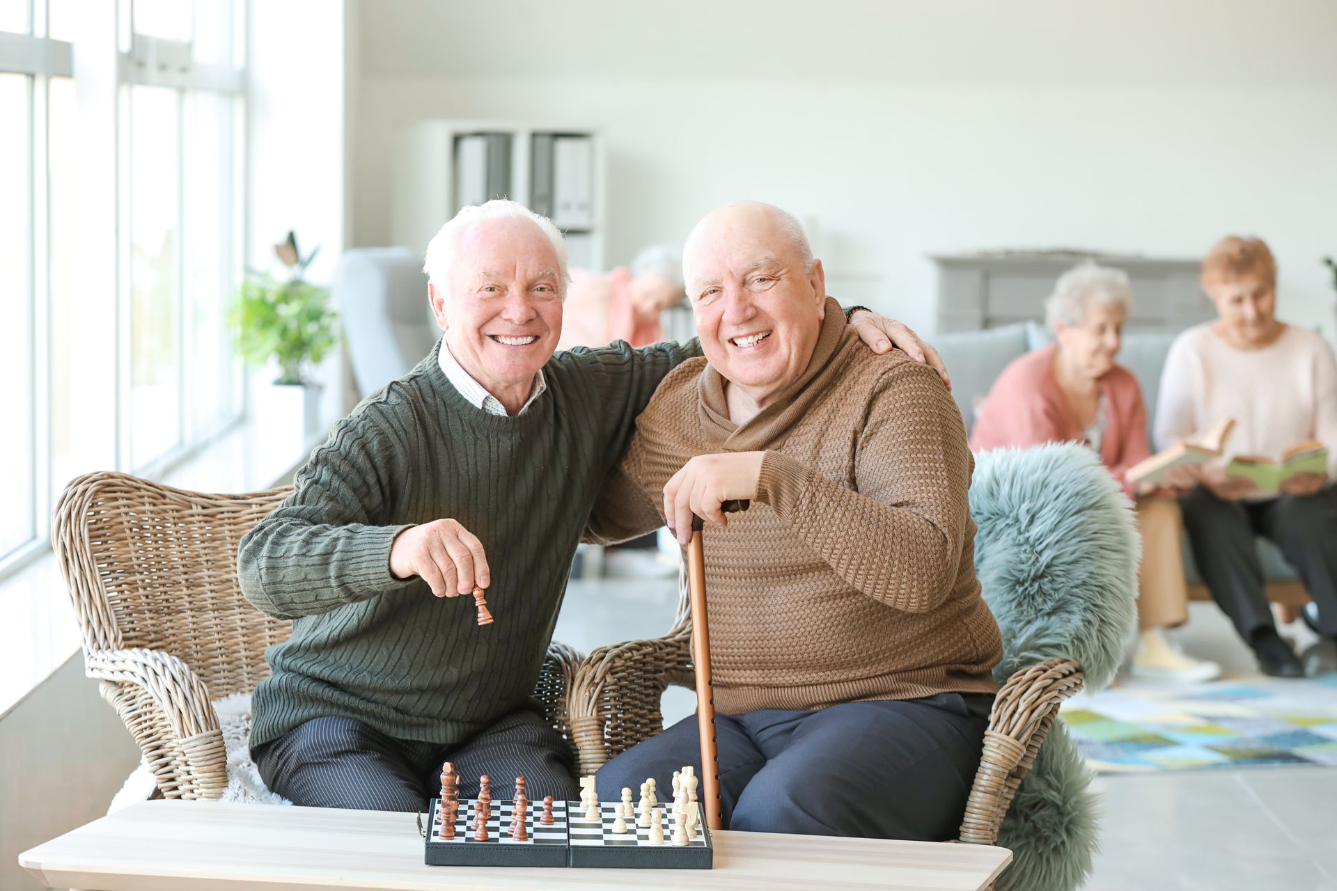 Two older men playing chess, smiling, one with arm around the other. Others in background.