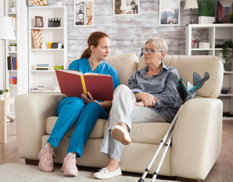 Nurse in blue scrubs reviews a book with an elderly person on a couch. Crutches are nearby.