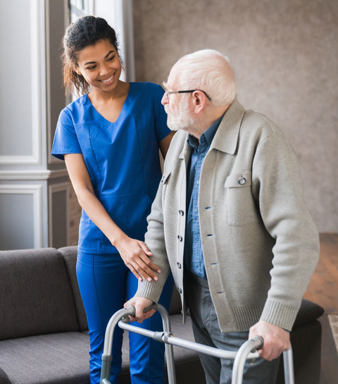 Caregiver in blue scrubs assists an older person using a walker in a home setting.