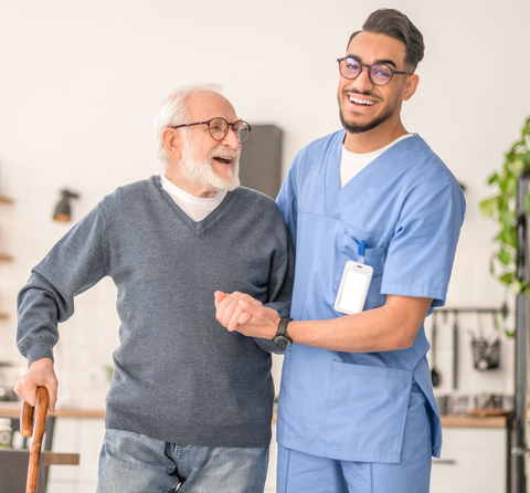 Caregiver helps elderly man walk with a cane, both smiling indoors.