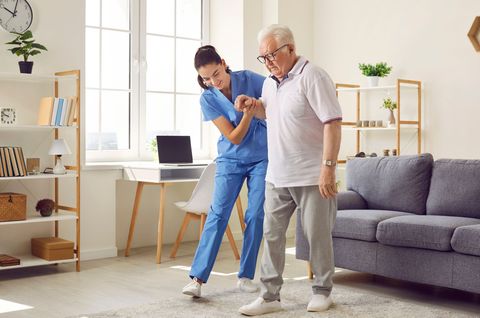 Nurse assists an older person walking in a living room, holding his arm. Sunlight streams in.