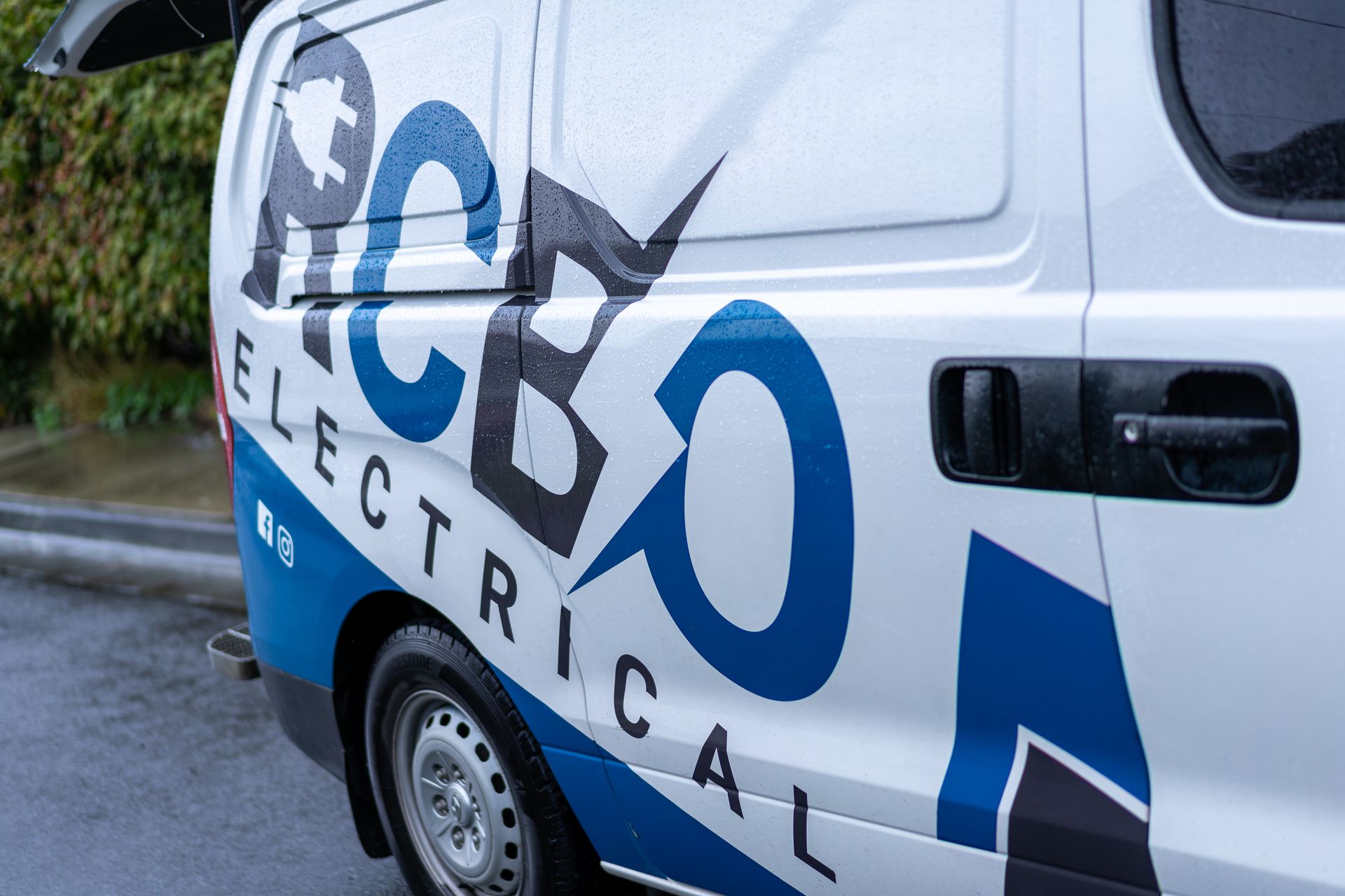White electrical van with blue accents and logo parked on wet street.