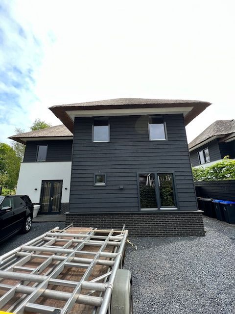 Huis met twee verdiepingen, donkergrijze gevelbekleding, witte muur en rieten dak. Er staat een stacaravan op de oprit.