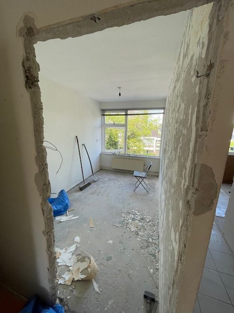 Kamer die gerenoveerd wordt, gezien door een deuropening. Puin op de vloer, onafgewerkte muren, raam op de achtergrond.