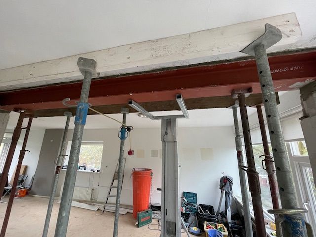 Bouwterrein met tijdelijke steunbalken die een rode stalen balk en een betonnen plafond ondersteunen.