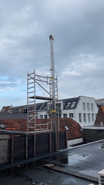 Steigers ondersteunen een hoge metalen schoorsteen op een dak, met een bewolkte lucht op de achtergrond.