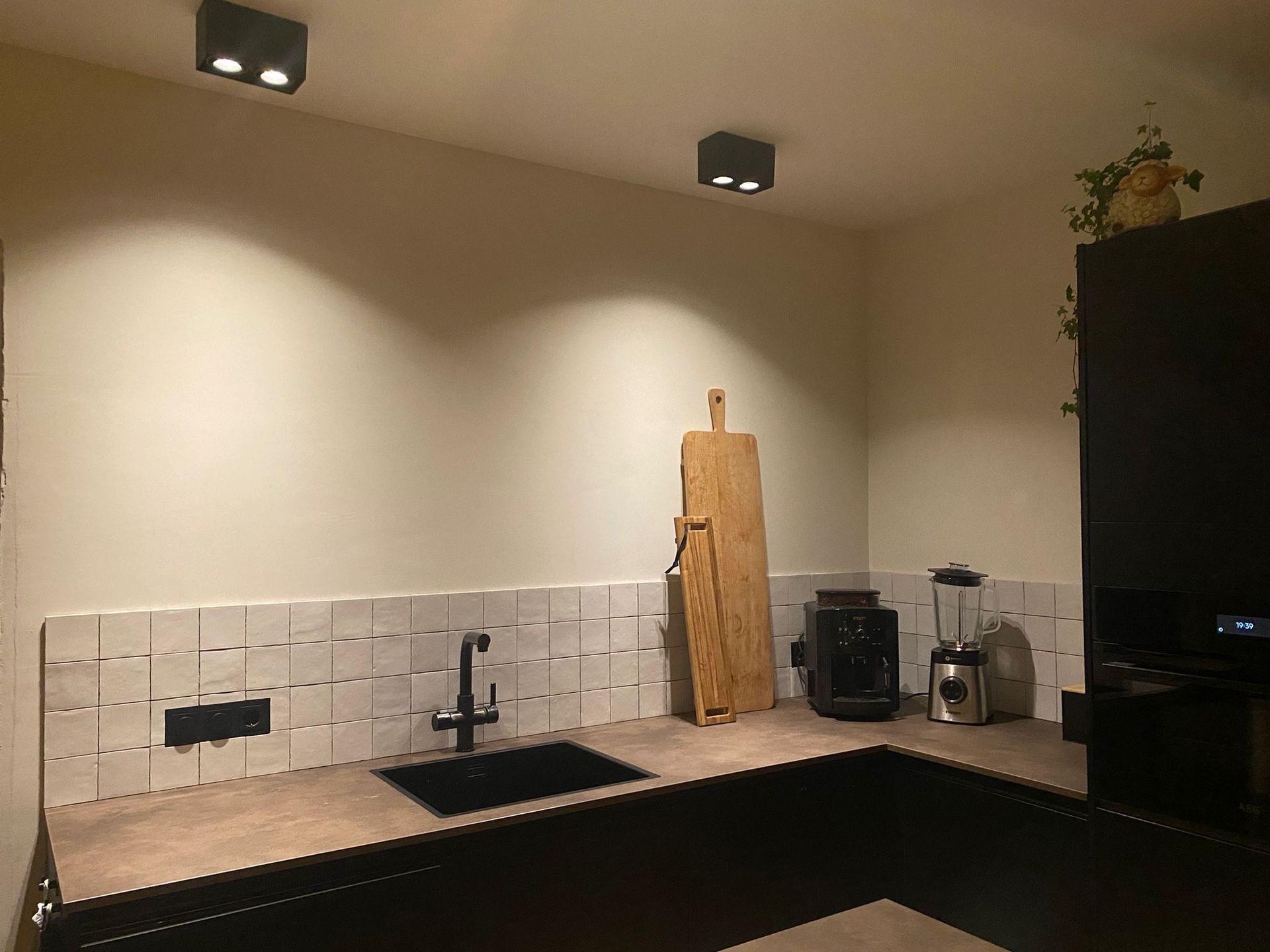 Keukenblad met spoelbak, apparatuur en houten snijplank. Donkere kasten, witte tegelachterwand en plafondverlichting.