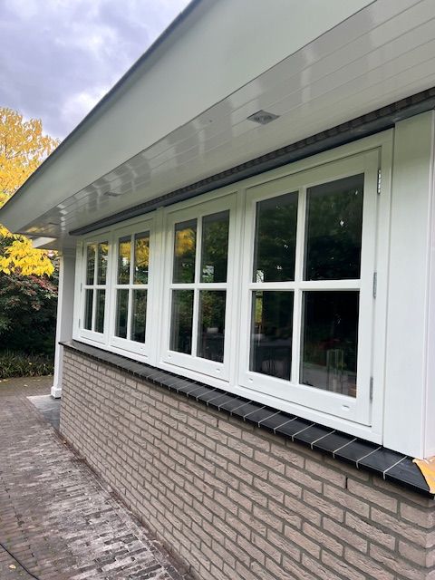 Witte ramen langs een bakstenen muur, onder een witte overstek. Zonlicht weerkaatst op de ramen.