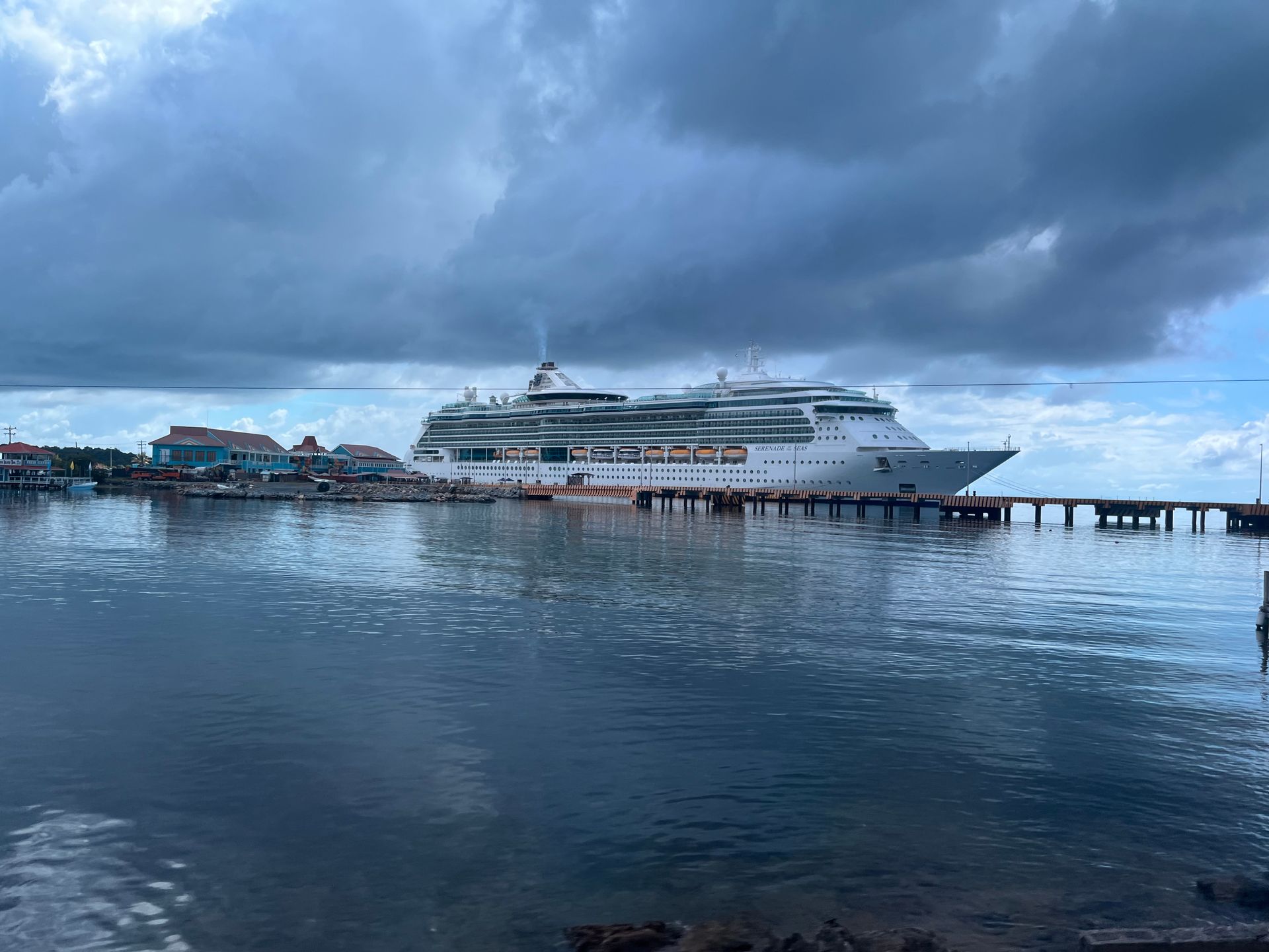 Cruise ship docked at a pier under a cloudy sky. Buildings line the port. Calm water in foreground.