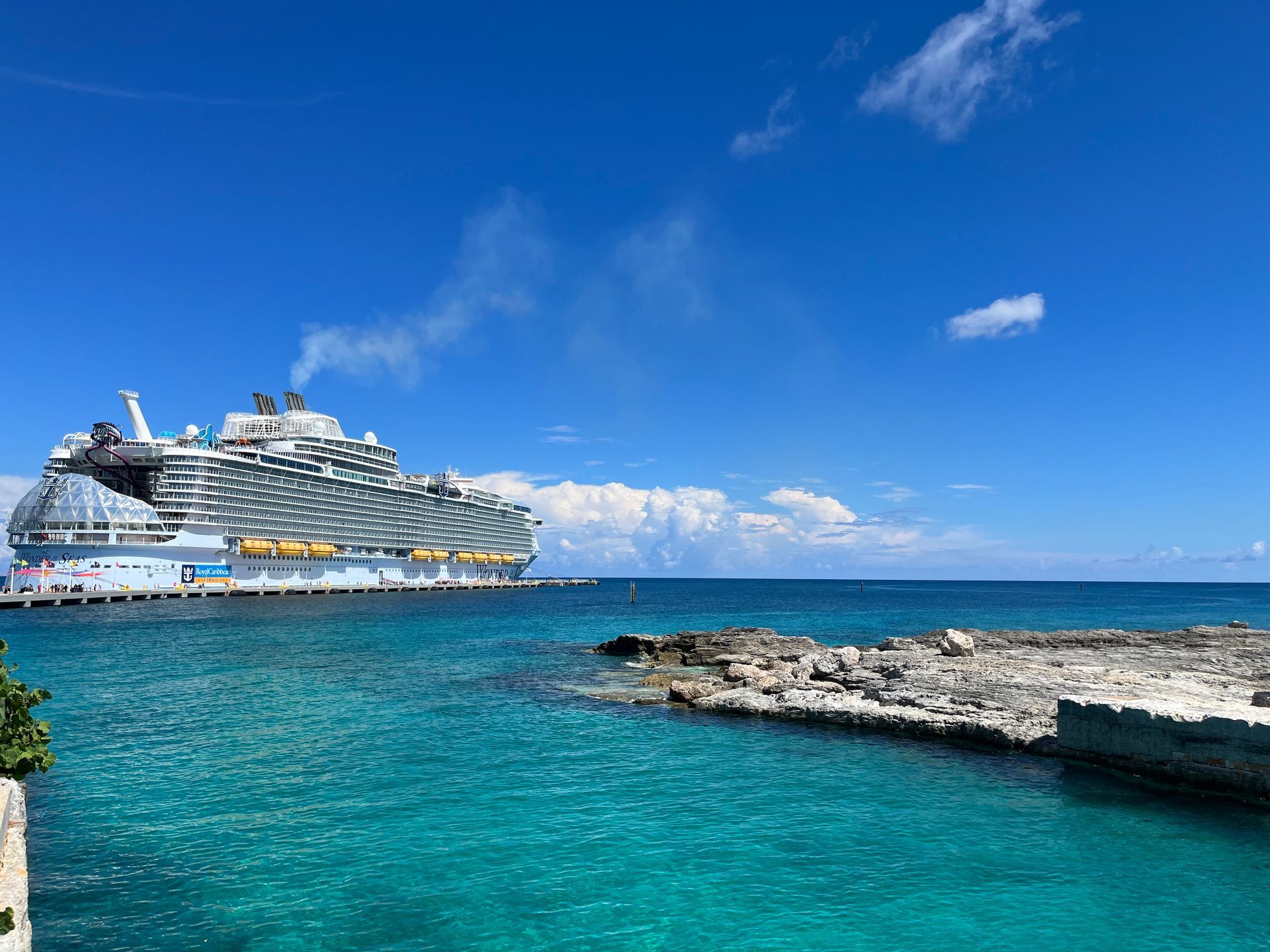 Cruise ship docked near a rocky shoreline on a sunny day with clear turquoise water.