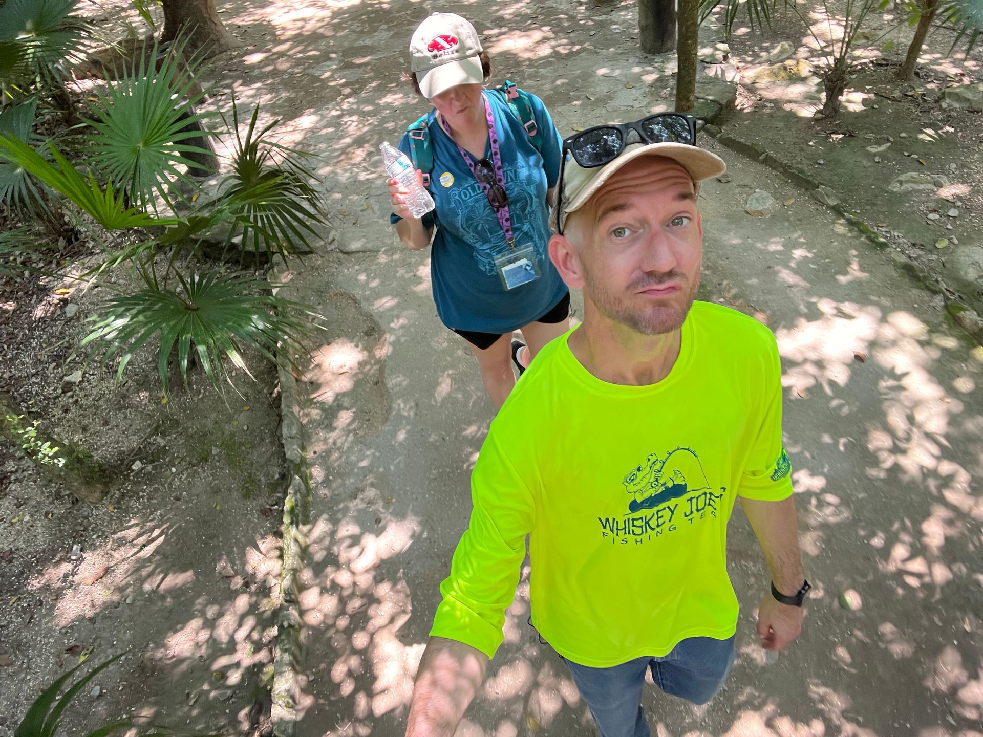 Man in neon yellow shirt takes selfie with woman behind him on a path, trees overhead.