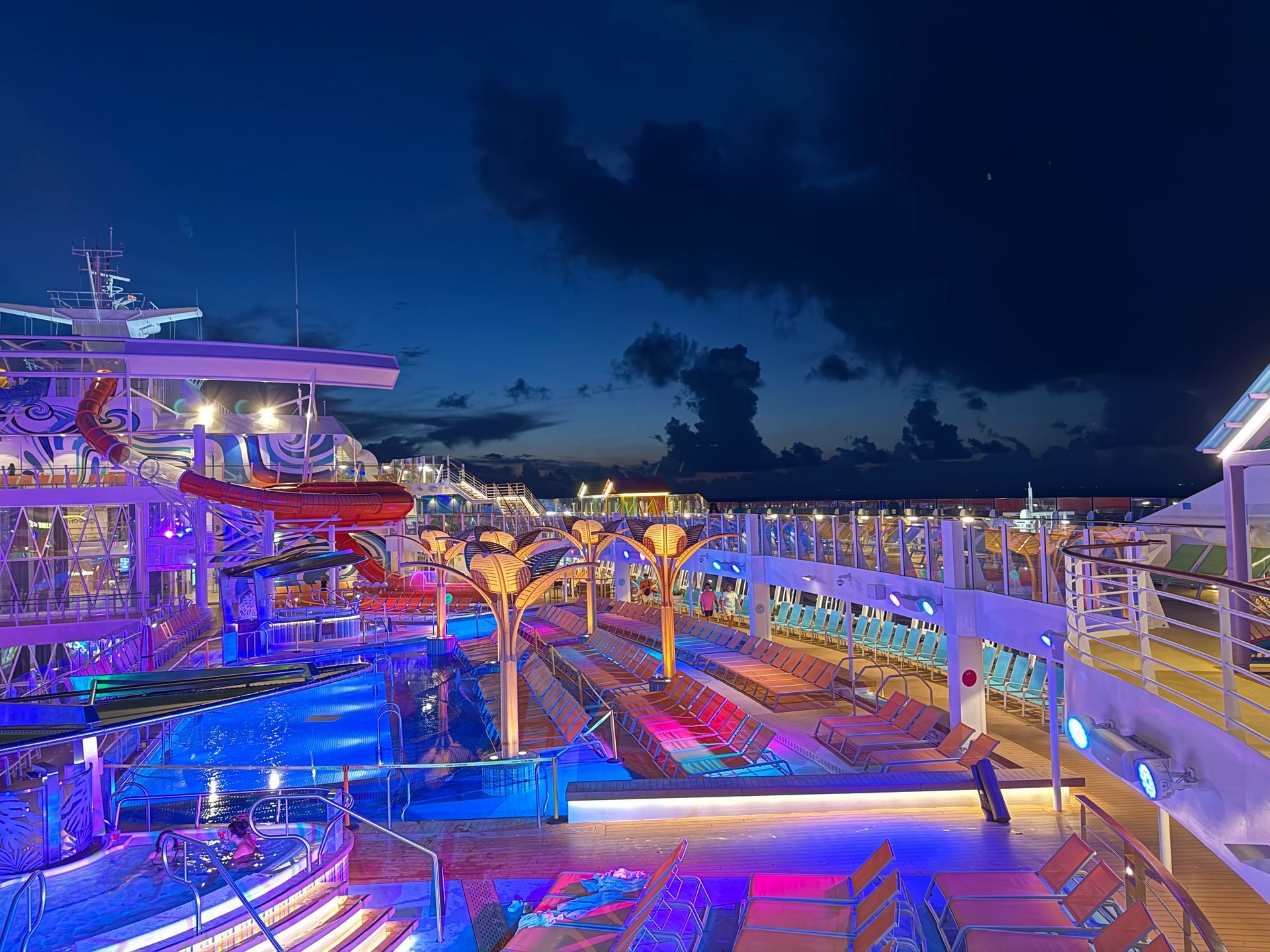Pool deck on a cruise ship at night, illuminated with colorful lights.