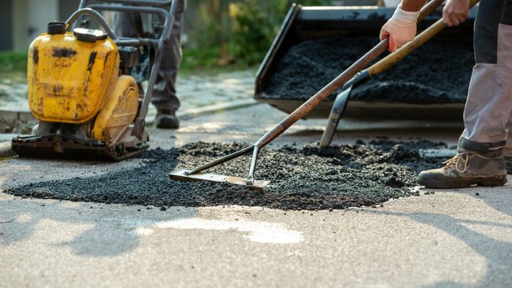 two people working with asphalt rakes to repair parking lot. Yellow construction machine in the background