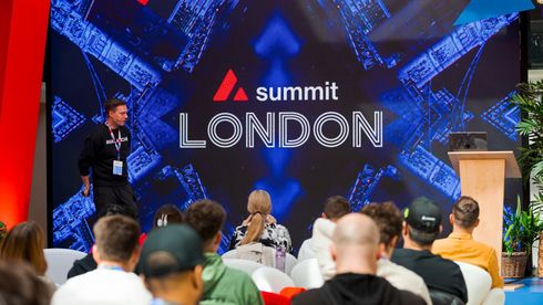 A speaker stands before an audience at the Atlassian Summit London, presented against a large blue, branded digital screen.