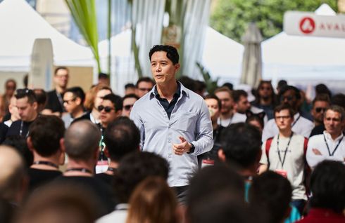 A person in a light blue shirt speaks to a crowded, blurry audience at an outdoor conference.