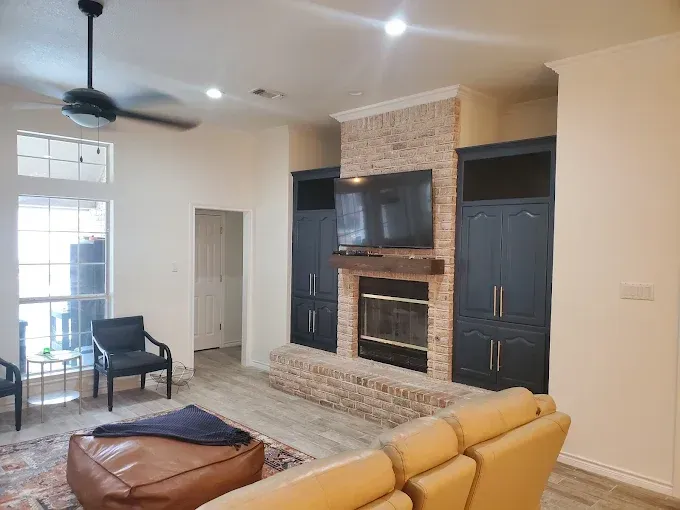 A living room featuring a tan leather sofa, an ottoman, and a brick fireplace flanked by dark blue built-in cabinets.