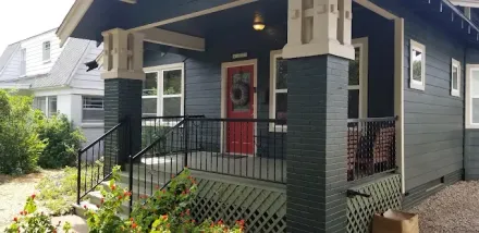 A dark gray house with a red front door, a porch with black railings, and brick pillars on a sunny day.
