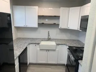 A modern kitchen featuring white cabinets, granite countertops, a farmhouse sink, and black appliances.
