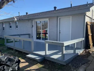 A gray painted wooden deck attached to the back of a single-story home with a sliding glass door and white door.