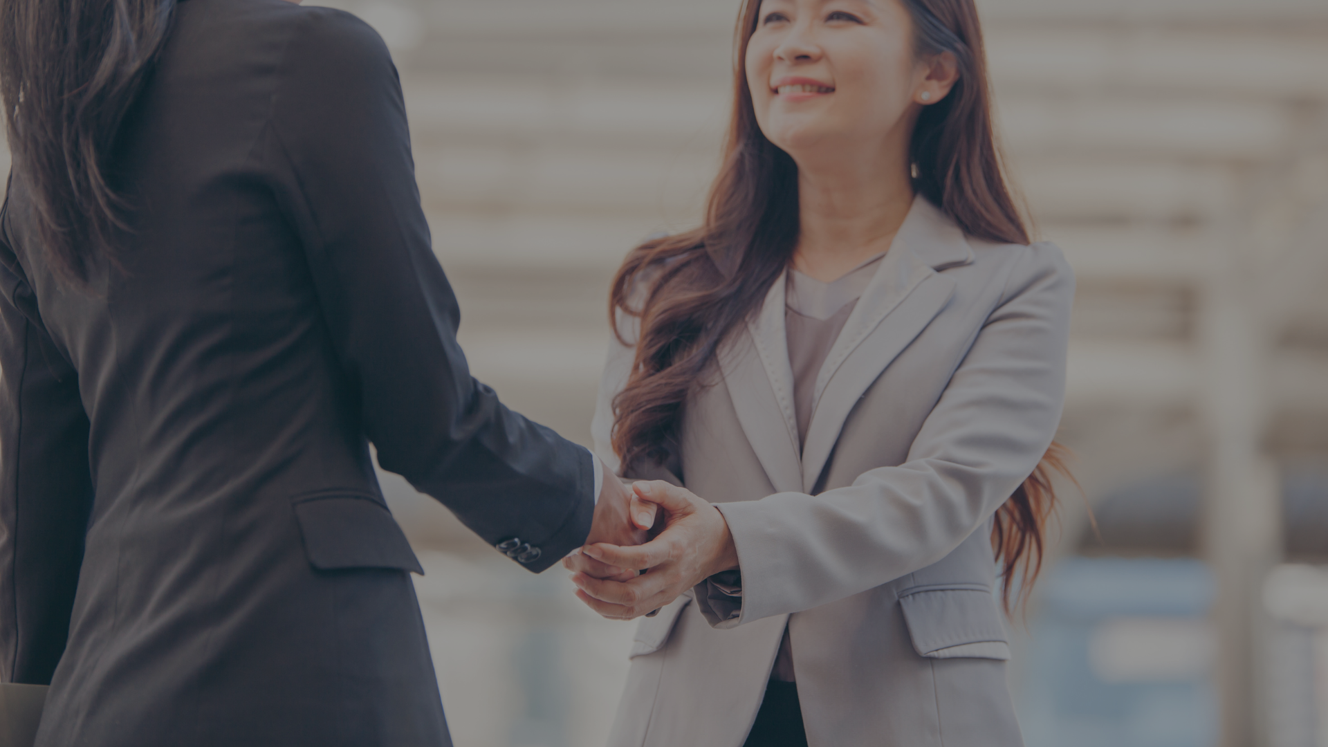 Two businesspeople shaking hands, smiling outdoors. One in a gray suit, the other in black.